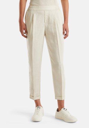 Osoba nosząca kremowe zwężane spodnie z podwiniętymi nogawkami i białe sportowe sneakersy na sznurowadła, stojąca na tle jasnego, jednolitego tła.