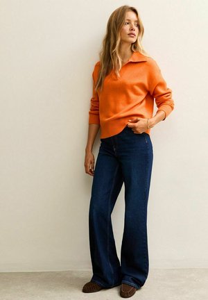 Oranje gebreide pullover trui met een kraag, gecombineerd met donkerblauwe flare jeans en bruine instap schoenen. Model poseert tegen een neutrale achtergrond.