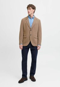 Brauner Blazer mit zwei Taschen, getragen über einem hellblauen Hemd. Dunkle Jeans und braune Schuhe vervollständigen das Outfit. Glatte Textur, strukturiertes Design.
