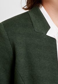 Tmavě zelený blazer s herringbone vzorem, s klopovým límcem a s jasně bílou košilí viditelnou pod ním.