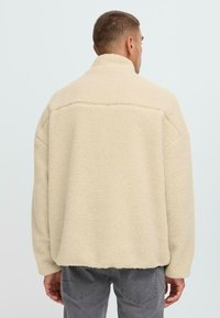 Beige Fleece-Jacke mit hohem Kragen, überschnittenen Schultern und strukturiertem Stoff. Hat einen geraden Saum und eine lockere Passform.