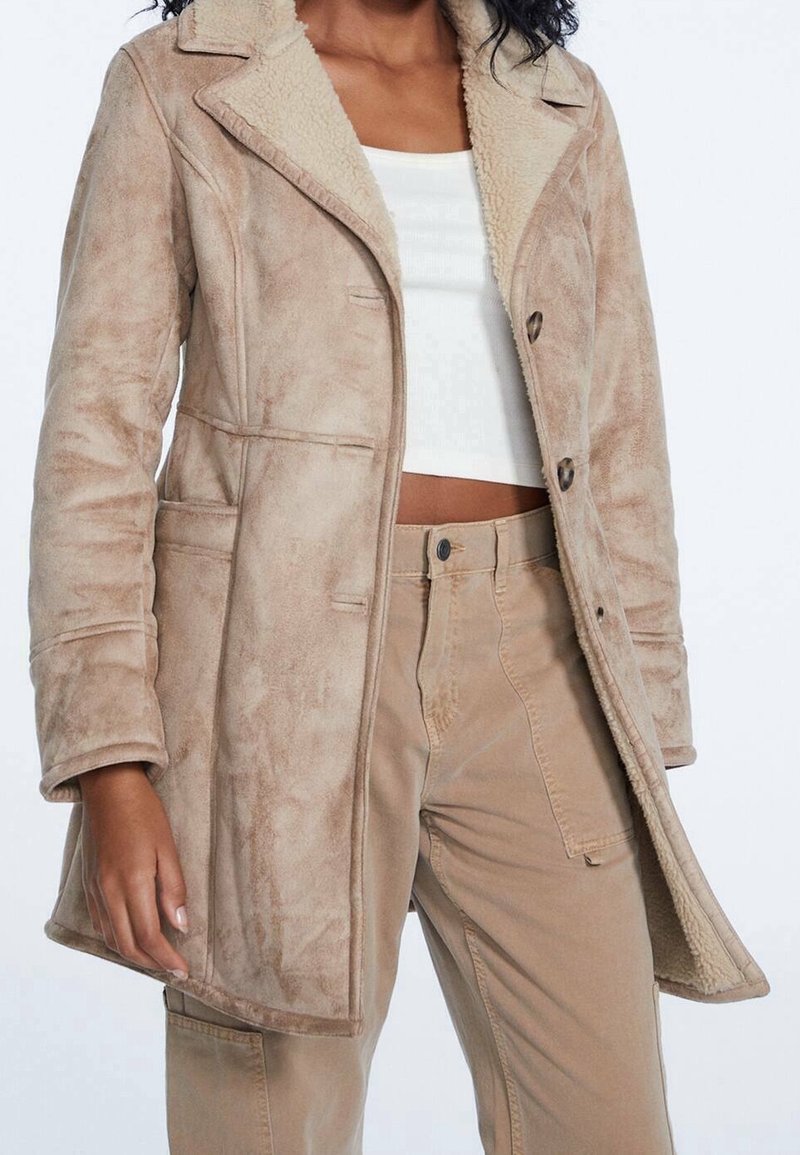 Beige nepbontjas met een op maat gemaakt ontwerp, voorzien van een opstaande kraag, knoopsluiting en zijzakken. Zachte textuur en warme voering.