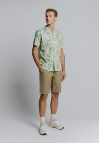 Patroon korte mouwen shirt in lichtgroen met abstracte designs, gecombineerd met zandkleurige shorts. Schoeisel bestaat uit witte sneakers en crew sokken.