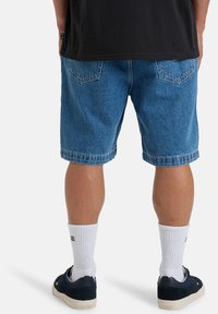 Denim-shortsit, sininen väri, löysä malli, käänteet lahkeissa. Valkoiset kommandosukat ja tummat kengät täydentävät asukokonaisuuden. Yksinkertainen musta paita näkyy yllä.