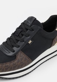 Baskets Michael Kors noir et marron en tissu texturé, lacets noirs, semelle blanche avec bande noire, et logo MK sur le côté et la languette.