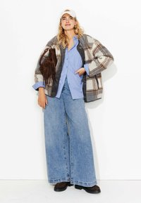 Veste en laine à carreaux gris et marron, chemise bleue à manches longues, jean en denim à jambe large, bottes en suède marron et casquette blanche. Sac à franges marron.