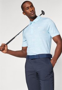Hellblaues Polohemd mit einem dezenten Muster, kurze Ärmel und einen Kragen. Kombiniert mit dunkelblauen Hosen und einem dunkelblauen Gürtel. Golfclub in der Hand gehalten.