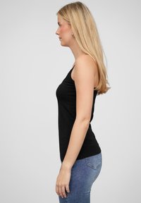 Svarte ermeless topp med en tettsittende design, laget av myk stoff. Vist iført med blå jeans i en profilvisning fra siden.