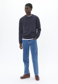 Felpa navy a girocollo, sotto un layer grigio chiaro, jeans blu a gamba dritta, e scarpe da barca marroni con dettagli in oro.