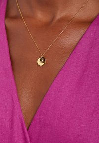 Goldkette mit einem runden Anhänger, der ein Sonnen- und Monddesign zeigt, auf einem tiefroten Stoffhintergrund getragen.