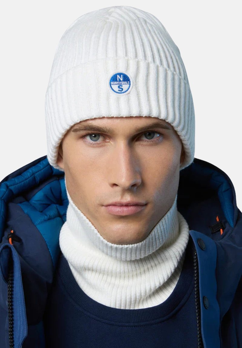 Berretto a coste bianco con logo circolare blu. Accompagnato da un dolcevita a coste bianche abbinato. Giacca imbottita blu visibile.