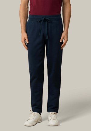 Hombre con pantalones deportivos azul marino con cordón y zapatillas blancas, de pie contra un fondo liso gris claro.