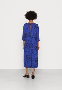 Vestido azul de mangas compridas com um ajuste solto, apresentando um padrão de respingos pretos e um detalhe de fenda na gola. Usado com mules pretas de salto alto.