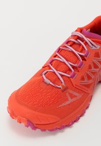 Sapatilha atlética laranja brilhante com parte superior em malha, atacadores cor-de-rosa, sobreposições estampadas e sola texturizada com cravos de tração roxos.