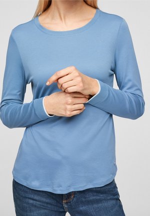 Vrouw die een lichtblauwe top met lange mouwen en donkerblauwe spijkerbroek draagt, met haar handen samengevouwen nabij de taille tegen een neutrale achtergrond.