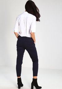 Witte, korte mouwen blouse met zijdoorsnedes, gecombineerd met navy, aansluitende broek. Model draagt zwarte, hoge enkellaarsjes. Achteraanzicht.