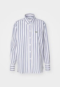 Chemise blanche à manches longues avec des rayures verticales bleues, une poche sur la poitrine et un logo brodé vert sur la poche.