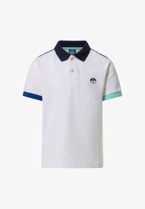 Garçon souriant portant un polo blanc avec des manches bleues et vertes, un short vert, des chaussettes blanches et des baskets blanches, se tenant devant un fond uni.