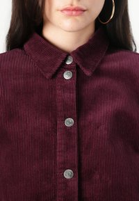 Bordeaux corduroy overhemd met een kraag, met verticale ribbels en zilveren drukknoopjes, dat een gestructureerd en samenhangend ontwerp laat zien.