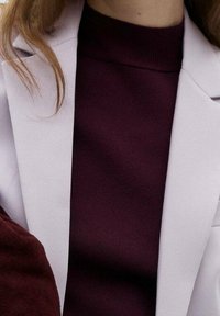 Col roulé bordeaux à texture côtelée, superposé sous un blazer cintré gris clair. Tissu lisse, coupe ajustée, sans pièces métalliques visibles.