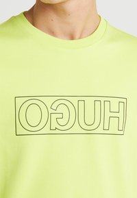 T-shirt gialla neon con logo "HUGO" contornato in nero in un design rettangolare. Collo rotondo, maniche corte, texture in cotone morbido.