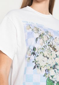 Bílý bavlněný tričko s modrým kostkovaným potiskem a květinovou zátiší designem ve zelené váze, s texturovanými květinovými detaily a krátkými rukávy.