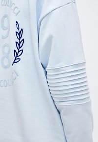 Γαλάζιο πουκάμισο μακρυμάνικο με ριμπ λεπτομέρεια στους αγκώνες και κεντημένο αριθμό «1988» και σχέδιο φύλλου στο πάνω μέρος του μπράτσου και στο στήθος.