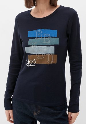 Femme portant un t-shirt à manches longues bleu marine avec des rayures colorées et le texte « BE YOU you got this! » sur le devant.