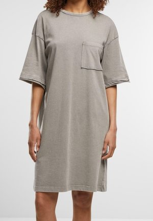 Frau trägt ein locker sitzendes, knielanges, hellgraues T-Shirt-Kleid mit kurzen Ärmeln und einer einzelnen Brusttasche vor schlichtem weißem Hintergrund.