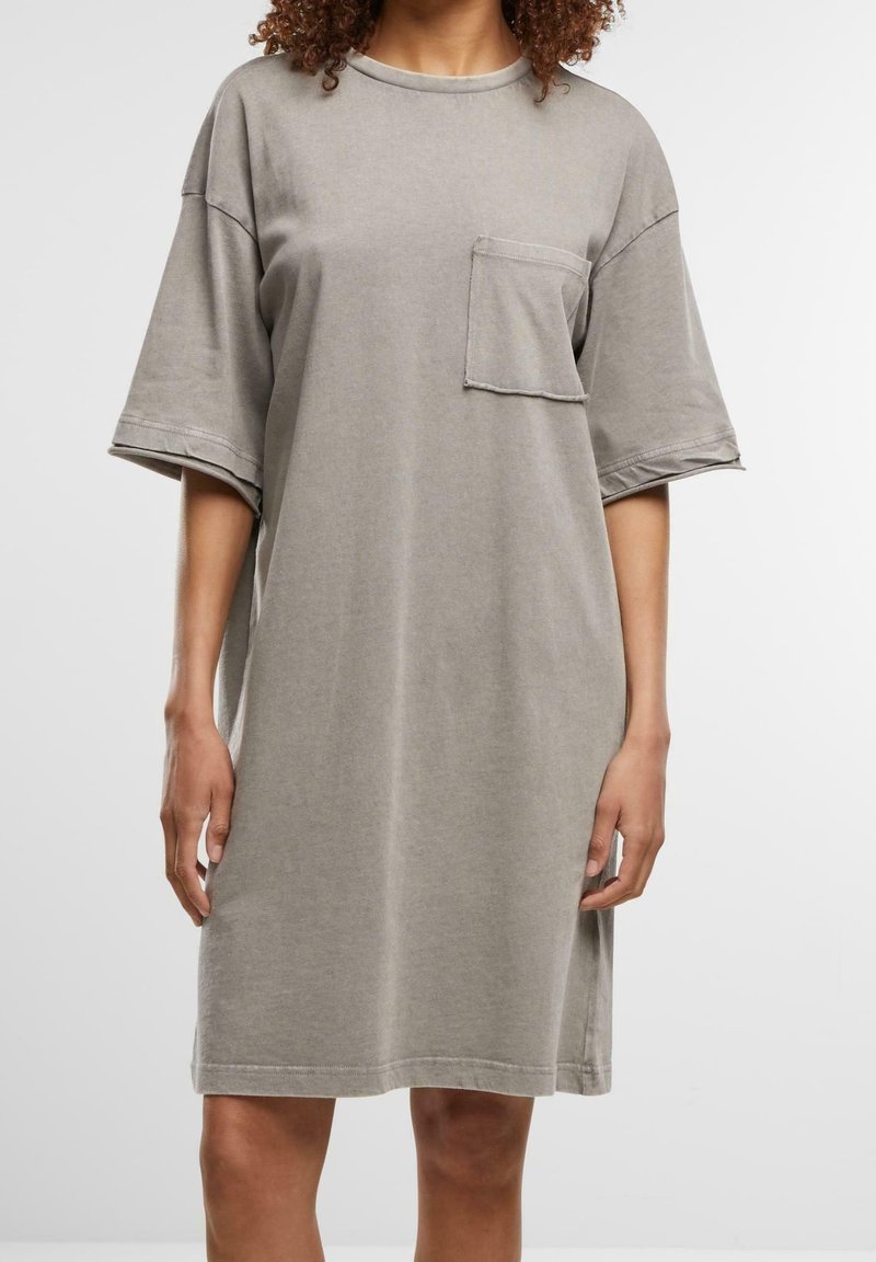 Femme portant une robe T-shirt ample, gris clair, longueur genou, à manches courtes et une seule poche poitrine, sur fond blanc uni.