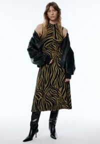 Gestreiftes Kleid in Olivgrün und Schwarz, ärmelloses Design, kombiniert mit einer übergroßen schwarzen Lederjacke und kniehohen schwarzen Stiefeln.