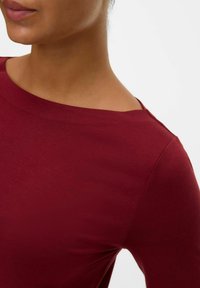Bordeaux top met een gladde textuur, ruime halslijn en lange mouwen, met een naaddetail langs de schouder.