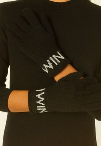 Guanti neri lavorati a maglia con una texture a coste, dotati di lettere ricamate grigie sui polsini. I guanti hanno un design senza dita.