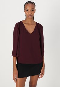 Bordo bluza s V-izrezom i rukavima do tri četvrtine, izrađena od lagane tkanine. Glatka tekstura, blago labav kroj, uparen s tamnom suknjom.