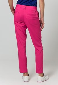 Osoba stojąca, ubrana w jaskraworóżowe spodnie, niebieską bluzkę i białe trampki, widziana od tyłu na jednolitym tle.