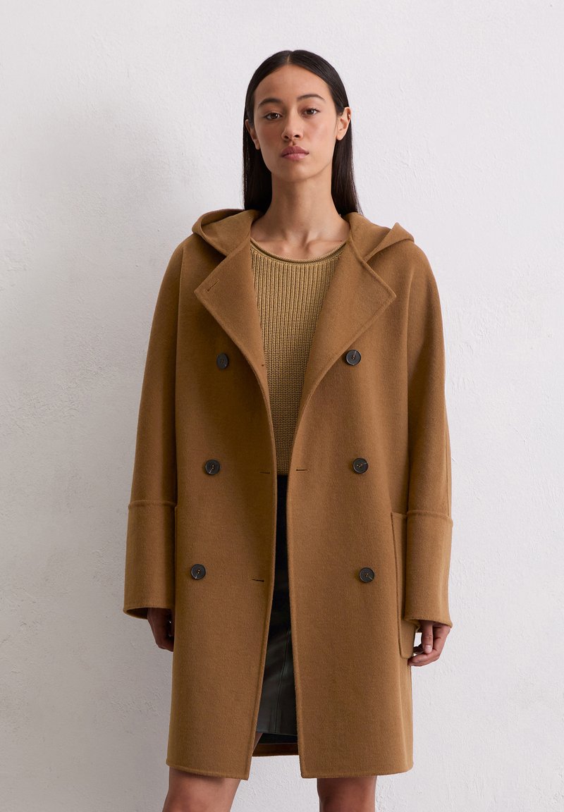 Manteau en laine marron avec capuche, design croisé, grands boutons noirs et manches surdimensionnées, porté sur un haut en tricot.