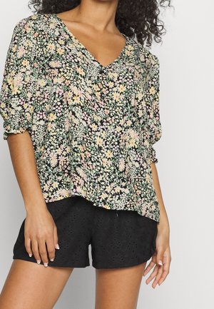 Kobieta w kwiecistej bluzce z dekoltem w serek, z bufkami na rękawach oraz w czarnych szortach z ażurowymi wycięciami, stojąca na tle jednolitego jasnego tła.