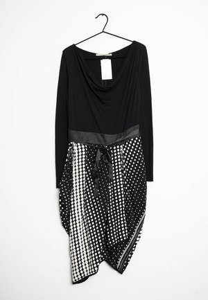 Schwarzes Langarmkleid mit einem drapierten Ausschnitt, das auf schwarzem Stoff ein weißes Punktmuster aufweist und in der Taille mit einem Satinband gebunden ist.