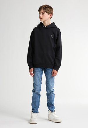Zwarte sweatervest met capuchon in een relaxte pasvorm, met ribgebreide manchetten en een klein rond logo op de borst, gecombineerd met lichtblauwe jeans en witte sneakers.