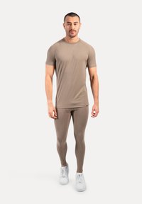 Man staand, met het gezicht naar voren, in een bijpassende taupe sportset met een T-shirt met korte mouwen, leggings en witte sneakers op een witte achtergrond.