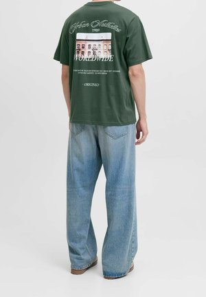Mężczyzna w ciemnozielonym graficznym T-shircie z obrazkiem budynku i napisem, w połączeniu z luźnymi jasnoniebieskimi dżinsami i brązowymi butami, stojący na białym tle.