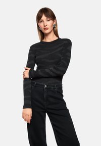 Black ribbed sweater with a subtle camo pattern; fitted design, long sleeves. Paired with high-waisted black jeans, featuring a button closure.