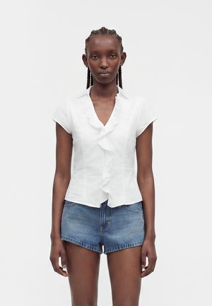Kvinne med flettehår som har på seg en hvit bluse med korte ermer og volanger og blå jeansshorts, stående foran en ensfarget lys bakgrunn.