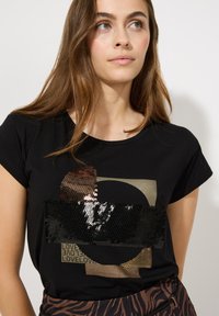Junge Frau mit langen braunen Haaren, die ein schwarzes T-Shirt trägt, das mit geometrischen Paillettenmustern und dem wiederholten Wort "LIEBE" bedruckt ist.