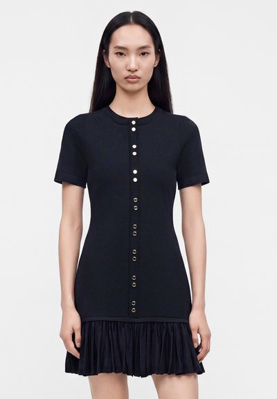 Femme aux longs cheveux noirs porte une robe noire ajustée avec des manches courtes, des boutons à l'avant et un ourlet plissé sur un fond clair uni.