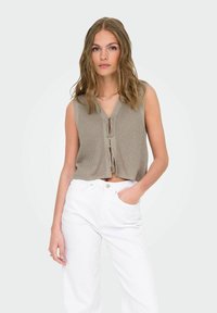 Strik, ærmeløs cropped top i lys taupe med V-udskæring, featuring en front knaplukning og ribbet tekstur. Parret med hvide jeans.