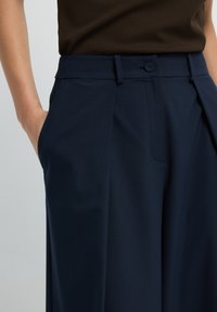 Donna che indossa pantaloni sartoriali blu navy con pieghe, una mano in tasca e un top senza maniche marrone aderente.