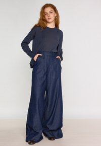Pantalons en denim à jambes larges bleu foncé avec une taille haute, dotés de plis avant et de poches latérales, associés à un haut ajusté bleu marine et un cardigan.