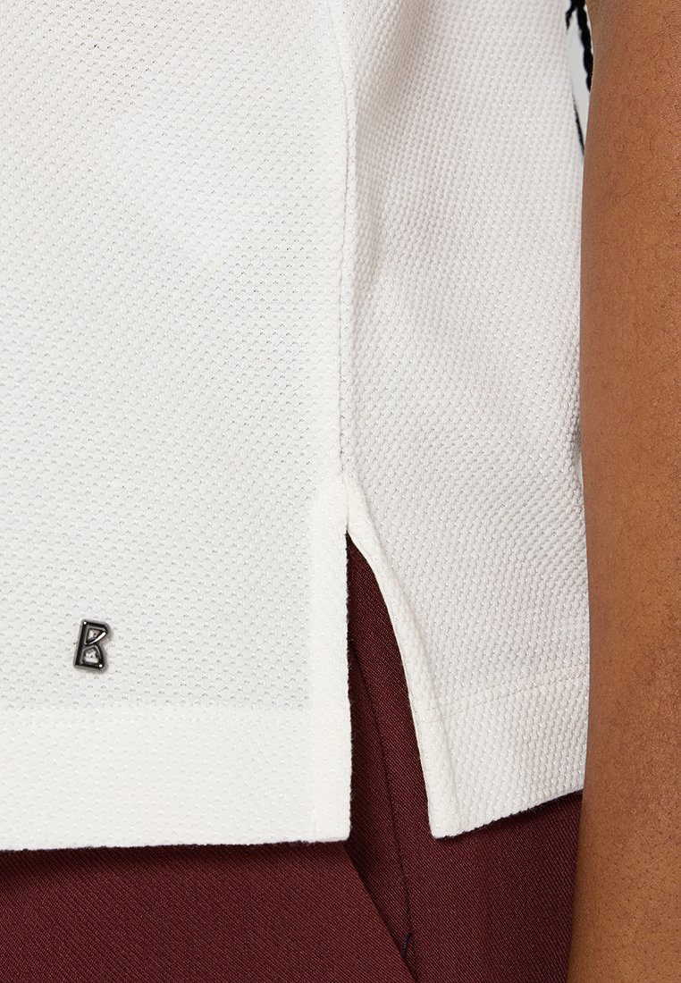 Haut blanc texturé avec une fente sur le côté, arborant un petit accent de logo métallique. Le tissu est respirant et il est associé à un bas de couleur bordeaux foncé.