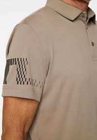 Mężczyzna ubrany w beżową, krótkorękawową koszulkę polo z czarnym pikselowym wzorem na górnej części lewej rękawki, kołnierzyk zapięty na guziki.